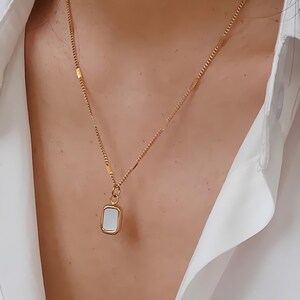 May include: Gold-toned necklace with a rectangular pendant. The pendant features a white, rectangular inlay. The necklace is worn over a white collared shirt. The chain has a delicate design.