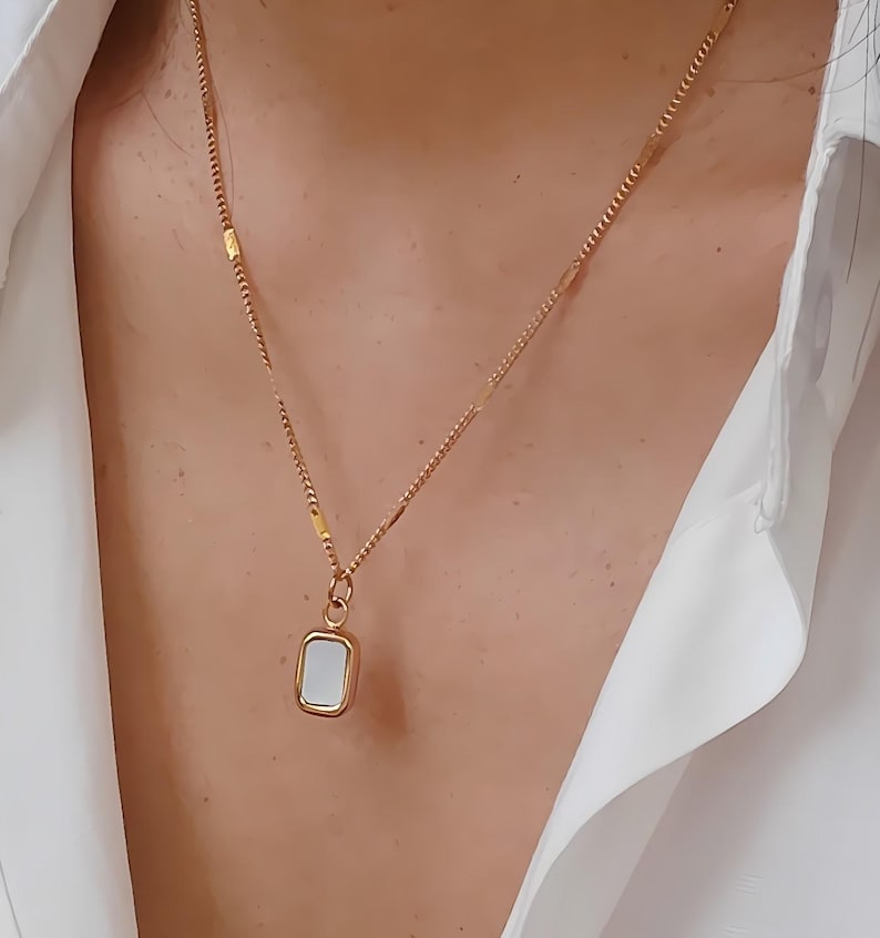 May include: Gold-toned necklace with a rectangular pendant. The pendant features a white, rectangular inlay. The necklace is worn over a white collared shirt. The chain has a delicate design.
