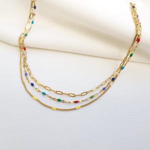 May include: A layered gold necklace with three strands. The top strand is a delicate chain, the middle has a paperclip chain, and the bottom has a chain with colorful beads. The necklace is displayed on a white surface.