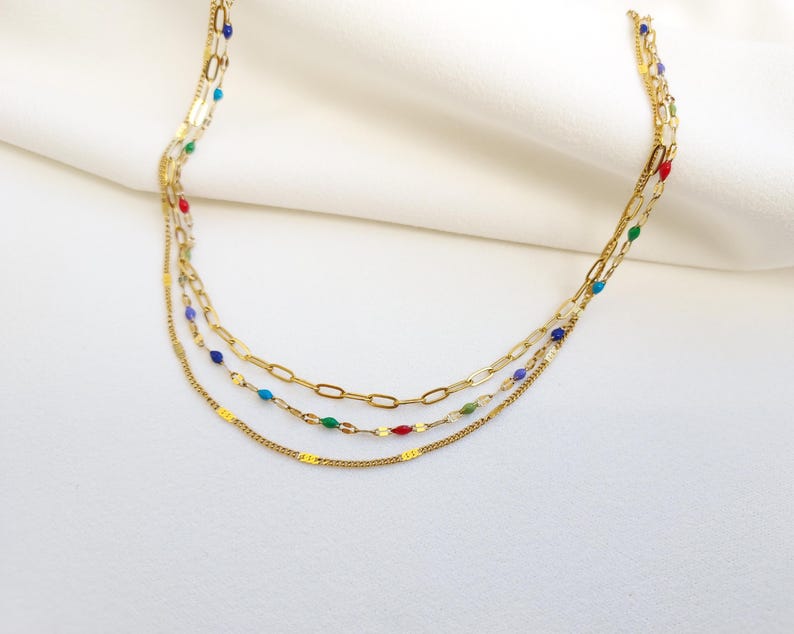 May include: A layered gold necklace with three strands. The top strand is a delicate chain, the middle has a paperclip chain, and the bottom has a chain with colorful beads. The necklace is displayed on a white surface.