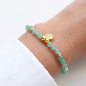 K&ouml;nnte beinhalten: Ein zartes Armband mit kleinen, runden, hellgr&uuml;nen Perlen, die mit goldfarbenen Perlen und Akzenten durchsetzt sind. Ein kleiner, runder, goldfarbener Anh&auml;nger h&auml;ngt am Armband. Das Armband wird am Handgelenk getragen, vor wei&szlig;em Hintergrund.