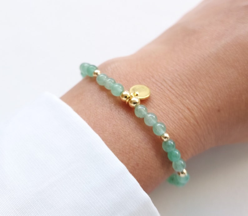 K&ouml;nnte beinhalten: Ein zartes Armband mit kleinen, runden, hellgr&uuml;nen Perlen, die mit goldfarbenen Perlen und Akzenten durchsetzt sind. Ein kleiner, runder, goldfarbener Anh&auml;nger h&auml;ngt am Armband. Das Armband wird am Handgelenk getragen, vor wei&szlig;em Hintergrund.