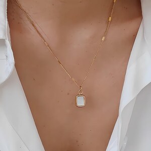 May include: A gold-colored necklace with a rectangular pendant. The pendant has a white, rectangular center and a gold-colored frame. The necklace is worn against a white shirt.