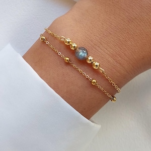 Peut inclure: Un bracelet en chaîne dorée avec une perle de pierre bleue et de petites perles dorées. Le bracelet est superposé à un bracelet en chaîne similaire avec de petites perles dorées.