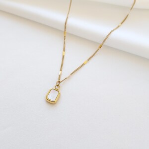 May include: Gold-toned necklace with a rectangular pendant. The pendant has a white, iridescent center and a gold frame. The chain is delicate, with a mix of small chain links and rectangular accents. The necklace is displayed on a white background.