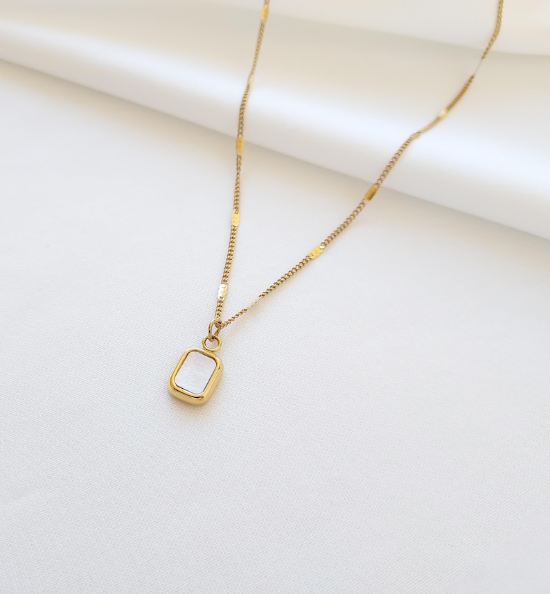 May include: Gold-toned necklace with a rectangular pendant. The pendant has a white, iridescent center and a gold frame. The chain is delicate, with a mix of small chain links and rectangular accents. The necklace is displayed on a white background.