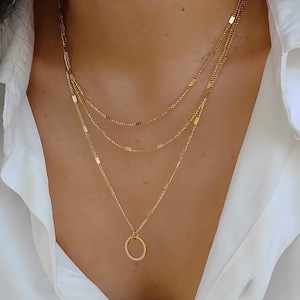 Peut inclure: Un collier superposé doré composé de trois chaînes délicates avec de petits accents métalliques plats et rectangulaires espacés régulièrement. La chaîne la plus longue comporte un pendentif circulaire simple suspendu au centre.