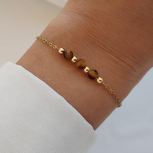 Op de afbeelding: Een delicate goudkleurige kettingarmband met drie gefacetteerde bruine tijgeroogkralen afgewisseld met vier kleine, gladde, ronde goudkleurige metalen kralen, gerangschikt in een enkele rij in het midden van de ketting.