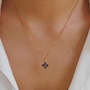 Könnte beinhalten: Goldfarbene Halskette mit einem vierblättrigen Anhänger. Jedes Blatt ist ein dunkelblauer Edelstein. Die Kette ist zart und hängt am Hals einer Person, vor einem weißen Hemd.