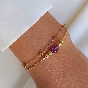 Peut inclure: Un bracelet en chaîne dorée avec une perle de pierre précieuse violette. Le bracelet est composé de deux chaînes superposées.