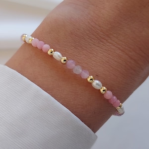 Peut inclure: Un bracelet délicat composé d'alternances de pierres roses, de perles blanches et de perles dorées. Le bracelet est monté sur un cordon fin et souple. Le bracelet est présenté sur un poignet, avec une chemise blanche en arrière-plan.