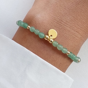 K&ouml;nnte beinhalten: Ein zartes Armband mit kleinen, runden, hellgr&uuml;nen Perlen, die mit goldfarbenen Akzenten versehen sind. Ein kleiner, runder, goldfarbener Anh&auml;nger h&auml;ngt am Armband. Das Armband wird am Handgelenk vor wei&szlig;em Hintergrund getragen.