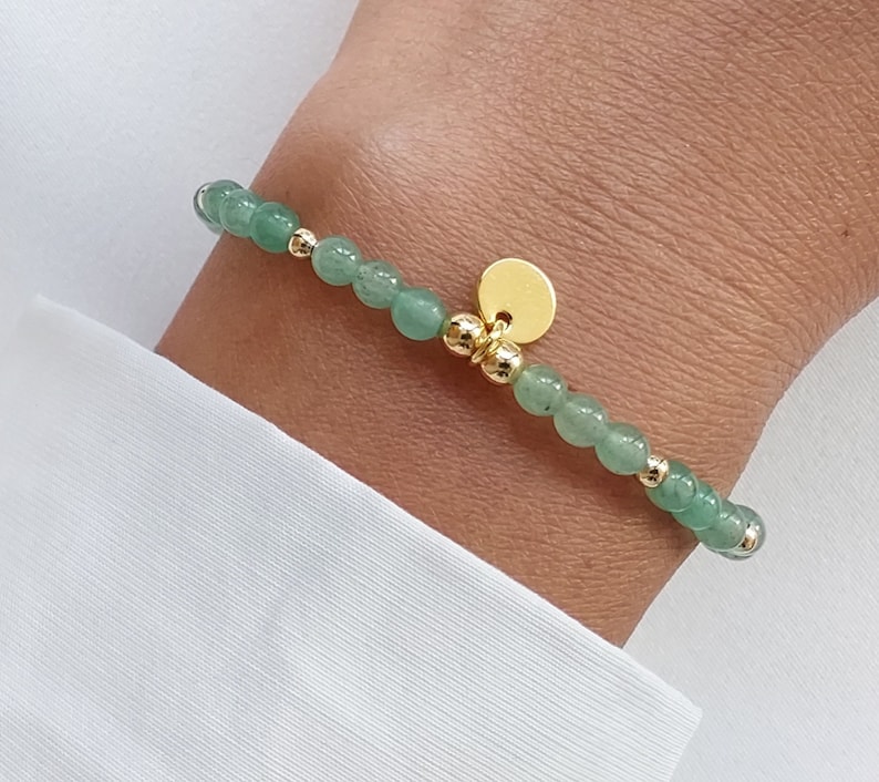 K&ouml;nnte beinhalten: Ein zartes Armband mit kleinen, runden, hellgr&uuml;nen Perlen, die mit goldfarbenen Akzenten versehen sind. Ein kleiner, runder, goldfarbener Anh&auml;nger h&auml;ngt am Armband. Das Armband wird am Handgelenk vor wei&szlig;em Hintergrund getragen.