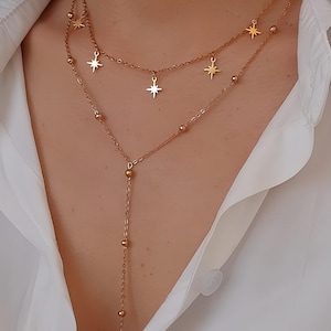 Puede incluir: Collar dorado en capas con colgantes de estrellas y cuentas. El collar tiene un diseño de cadena delicado con múltiples hilos, creando un efecto en capas. Las estrellas están espaciadas uniformemente, agregando un toque celestial a la joyería.