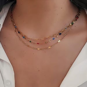 May include: A layered gold necklace set. The top chain features large oval links, while the middle chain has colorful beads. The bottom chain is a delicate, textured design. The necklaces are displayed against a white shirt.