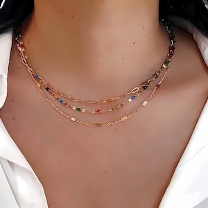 May include: A layered gold necklace with three strands. The top strand features a chain with colorful beads, the middle has a paperclip chain with colorful beads, and the bottom is a delicate chain.