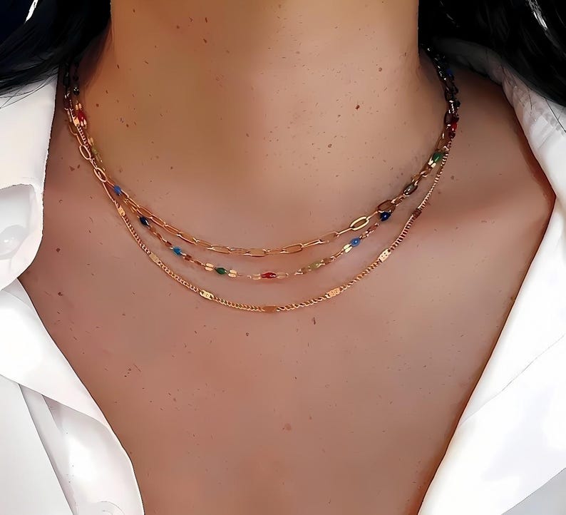 May include: A layered gold necklace with three strands. The top strand features a chain with colorful beads, the middle has a paperclip chain with colorful beads, and the bottom is a delicate chain.