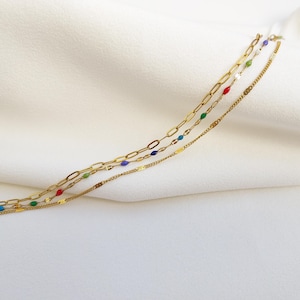 May include: Three gold necklaces with different chain styles. One chain has colorful enamel beads in red, green, blue, and white. The other two chains are simple gold link and flat chains. The necklaces are displayed on a white fabric background.