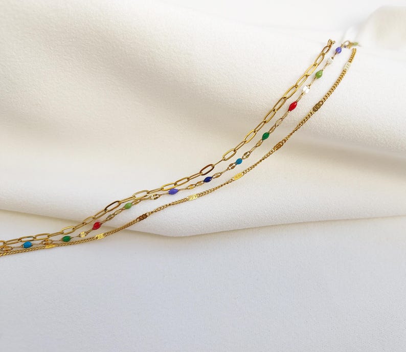 May include: Three gold necklaces with different chain styles. One chain has colorful enamel beads in red, green, blue, and white. The other two chains are simple gold link and flat chains. The necklaces are displayed on a white fabric background.