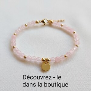 K&ouml;nnte beinhalten: Ein zartes Armband mit kleinen, hellrosa Rosenquarzperlen, die mit goldfarbenen Akzenten versehen sind. Ein kleiner, runder goldener Anh&auml;nger h&auml;ngt in der Mitte. Der Text "D&eacute;couvrez - le dans la boutique" steht unten.
