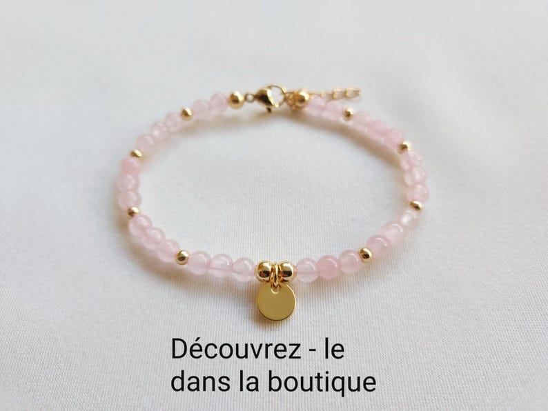 K&ouml;nnte beinhalten: Ein zartes Armband mit kleinen, hellrosa Rosenquarzperlen, die mit goldfarbenen Akzenten versehen sind. Ein kleiner, runder goldener Anh&auml;nger h&auml;ngt in der Mitte. Der Text "D&eacute;couvrez - le dans la boutique" steht unten.