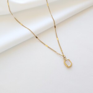 May include: A delicate gold-colored necklace with a rectangular pendant. The pendant has a light-colored, possibly mother-of-pearl, center stone. The chain has small, rectangular accents. The necklace is displayed on a white, textured surface.