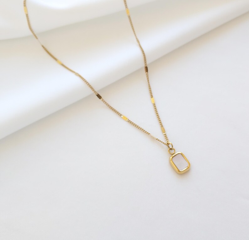 May include: A delicate gold-colored necklace with a rectangular pendant. The pendant has a light-colored, possibly mother-of-pearl, center stone. The chain has small, rectangular accents. The necklace is displayed on a white, textured surface.