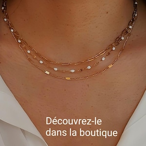 May include: A layered gold-toned necklace with three strands. The top strand features a delicate chain, the middle strand has white oval beads, and the bottom strand is a thin, textured chain. The text "D&eacute;couvrez-le dans la boutique" is visible.