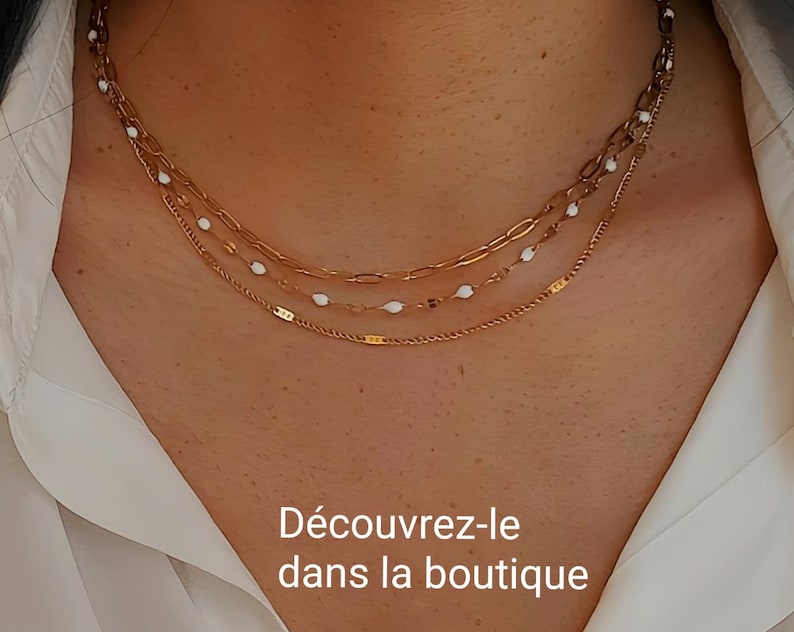 May include: A layered gold-toned necklace with three strands. The top strand features a delicate chain, the middle strand has white oval beads, and the bottom strand is a thin, textured chain. The text "D&eacute;couvrez-le dans la boutique" is visible.