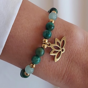 Peut inclure: Un bracelet de perles avec des pierres précieuses vertes et bleu clair, orné de perles dorées et d'un charm en forme de fleur de lotus dorée. Le bracelet est porté au poignet, mettant en valeur le design et la couleur du bijou.