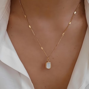 May include: A gold-colored necklace with a rectangular pendant. The pendant has a white, rectangular center and a gold border. The necklace is worn against a white shirt.