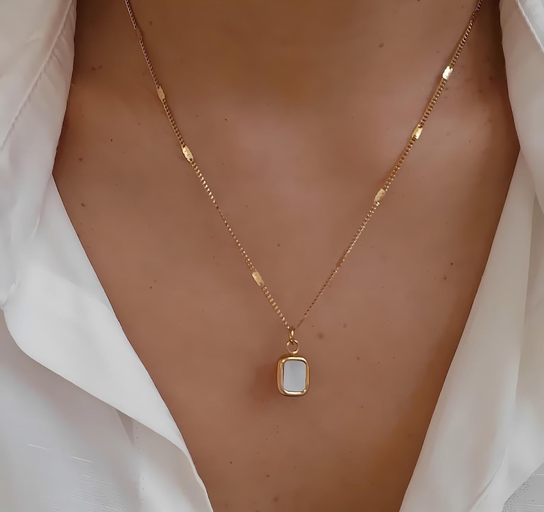 May include: A gold-colored necklace with a rectangular pendant. The pendant has a white, rectangular center and a gold border. The necklace is worn against a white shirt.