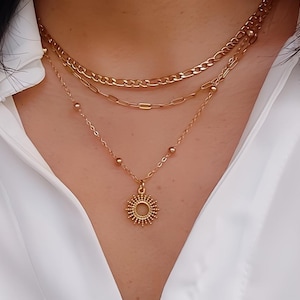 Könnte beinhalten: Eine mehrlagige goldene Halskette mit einem Sonnenanhänger. Die Halskette verfügt über drei verschiedene Kettenarten und einen zarten Sonnenanhänger.