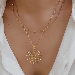 Peut inclure: Ensemble de colliers superposés dorés. La couche supérieure présente de petites perles, tandis que la couche inférieure comporte un pendentif avec un motif de soleil et un centre en forme de goutte. Les colliers sont portés sur une chemise blanche.