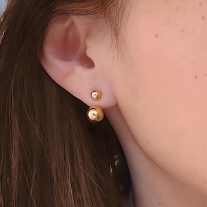 May include: A pair of gold-toned double-sided ball stud earrings. The earrings are worn in the earlobe.