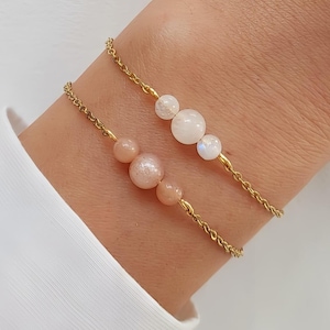 Op de afbeelding: Twee gouden kettingarmbanden met edelsteen kralen. De ene armband heeft drie witte maansteen kralen en de andere heeft drie perzikkleurige zonnesteen kralen.