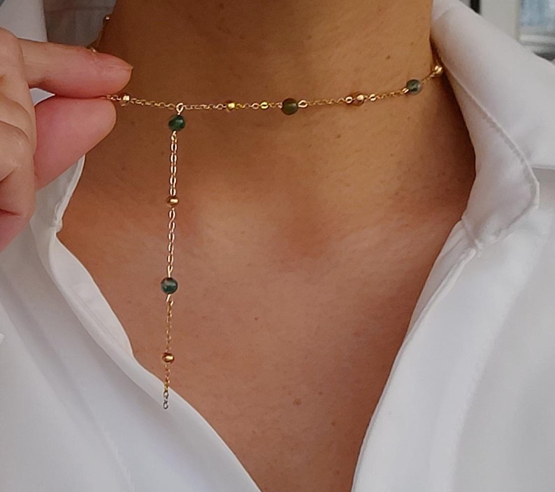 May include: A gold chain necklace with a delicate pendant featuring small green beads. The necklace is layered with a shorter chain that sits closer to the neck.
