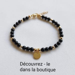 K&ouml;nnte beinhalten: Ein Armband mit schwarzen Perlen und goldfarbenen Elementen. Ein kleiner, runder goldener Anh&auml;nger h&auml;ngt in der Mitte. Das Armband wird auf einer wei&szlig;en Oberfl&auml;che pr&auml;sentiert. Der Text unten lautet: "Entdecke es im Shop."