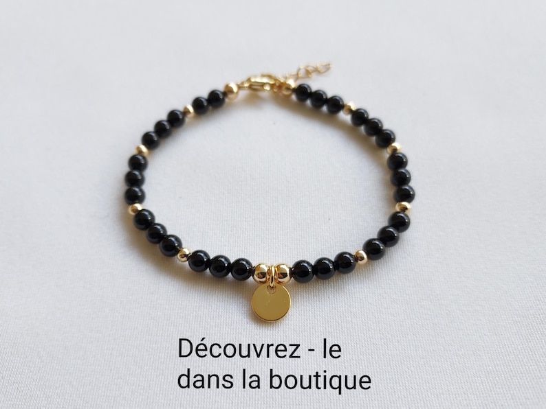 K&ouml;nnte beinhalten: Ein Armband mit schwarzen Perlen und goldfarbenen Elementen. Ein kleiner, runder goldener Anh&auml;nger h&auml;ngt in der Mitte. Das Armband wird auf einer wei&szlig;en Oberfl&auml;che pr&auml;sentiert. Der Text unten lautet: "Entdecke es im Shop."