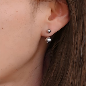 Peut inclure: Une paire de boucles d'oreilles boule argentées. Une petite boule est dans le lobe de l'oreille, et une plus grande boule est derrière le lobe. Design simple et moderne.
