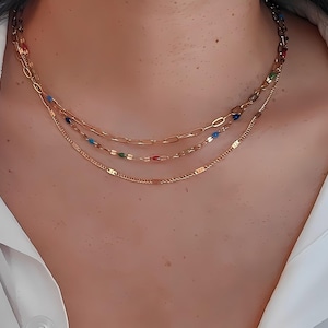 May include: A layered gold necklace with three strands. The top strand has a chain link design, the middle strand has colorful beads, and the bottom strand has a delicate chain. The necklace is worn against a white shirt.