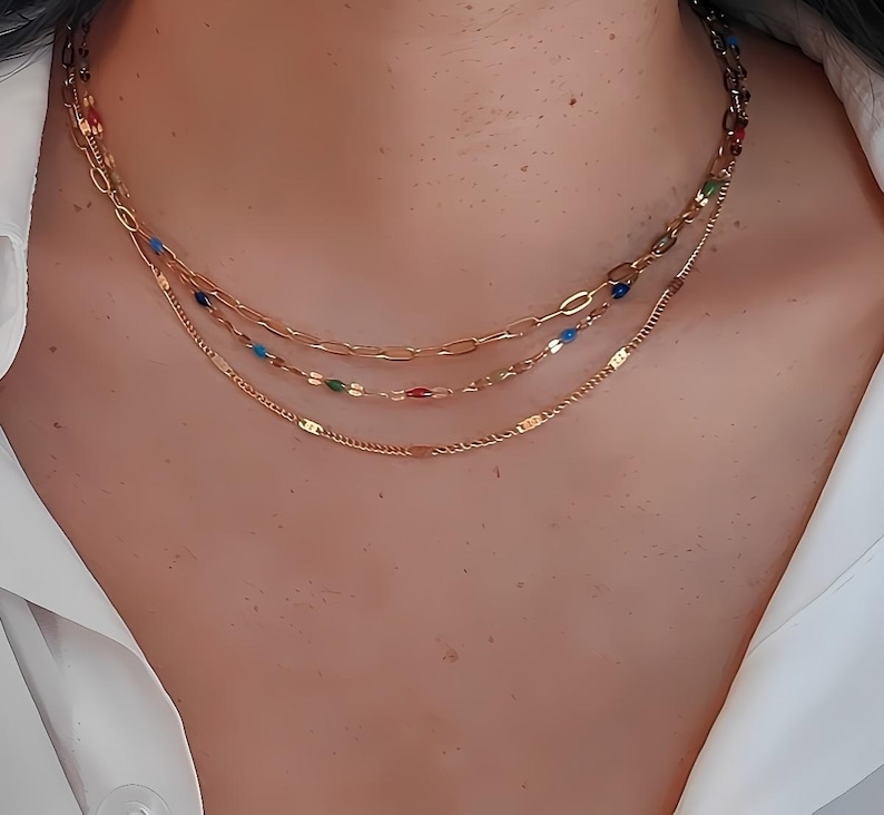 May include: A layered gold necklace with three strands. The top strand has a chain link design, the middle strand has colorful beads, and the bottom strand has a delicate chain. The necklace is worn against a white shirt.