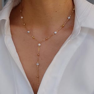 May include: A gold chain necklace with white beads and a long pendant. The necklace is layered and features small gold beads.
