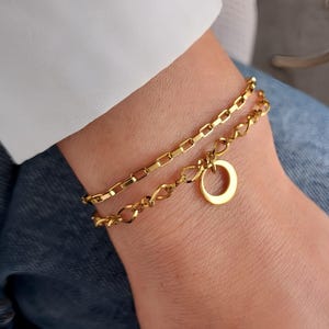 Peut inclure: Deux bracelets dorés sont portés sur un poignet. Le bracelet supérieur a des maillons rectangulaires, tandis que le bracelet inférieur a un design en chaîne avec un charme circulaire. Fond blanc et jean bleu.