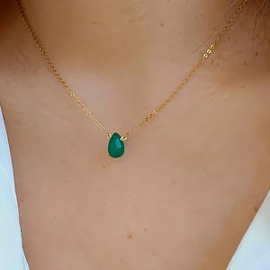 Puede incluir: Un collar dorado con un colgante de piedra preciosa verde esmeralda en forma de lágrima. El colgante cuelga de una delicada cadena, creando un diseño sencillo. El collar se lleva sobre una camisa blanca.