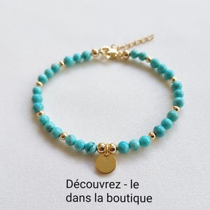 K&ouml;nnte beinhalten: T&uuml;rkisfarbenes Perlenarmband mit goldfarbenen Akzenten und einem kleinen, runden Anh&auml;nger. Das Armband hat kleine, runde t&uuml;rkisfarbene Perlen und goldfarbene Perlen. Der Verschluss und der Anh&auml;nger sind ebenfalls goldfarben. Der Text "D&eacute;couvrez - le dans la boutique" steht unten.