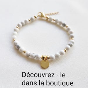 K&ouml;nnte beinhalten: Wei&szlig;-graues Perlenarmband mit goldenen Akzenten und einem goldenen Anh&auml;nger. Das Armband hat kleine, runde Perlen und eine zarte Kette. Der Anh&auml;nger ist eine kleine, runde, goldene Scheibe. Der Text "D&eacute;couvrez - le dans la boutique" ist sichtbar.