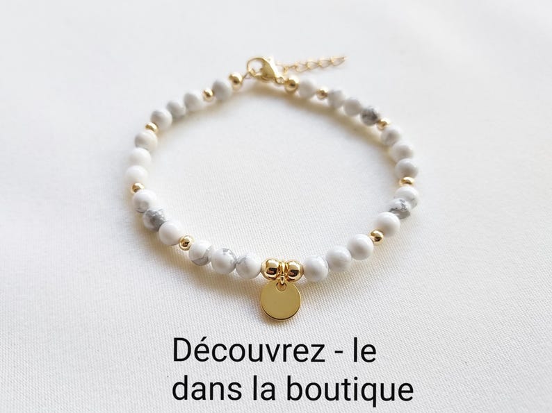 K&ouml;nnte beinhalten: Wei&szlig;-graues Perlenarmband mit goldenen Akzenten und einem goldenen Anh&auml;nger. Das Armband hat kleine, runde Perlen und eine zarte Kette. Der Anh&auml;nger ist eine kleine, runde, goldene Scheibe. Der Text "D&eacute;couvrez - le dans la boutique" ist sichtbar.