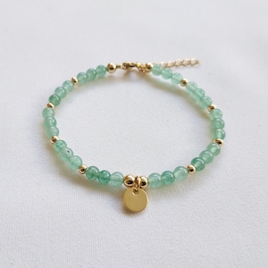 K&ouml;nnte beinhalten: Ein zartes Armband mit kleinen, hellgr&uuml;nen Perlen, die mit goldfarbenen Akzenten durchsetzt sind. Ein kleiner, runder goldener Anh&auml;nger h&auml;ngt in der Mitte. Das Armband wird auf einer wei&szlig;en Oberfl&auml;che pr&auml;sentiert.