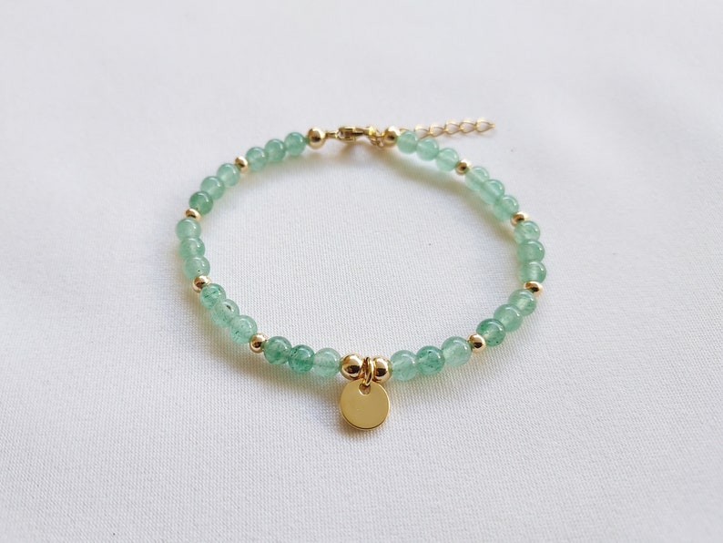K&ouml;nnte beinhalten: Ein zartes Armband mit kleinen, hellgr&uuml;nen Perlen, die mit goldfarbenen Akzenten durchsetzt sind. Ein kleiner, runder goldener Anh&auml;nger h&auml;ngt in der Mitte. Das Armband wird auf einer wei&szlig;en Oberfl&auml;che pr&auml;sentiert.